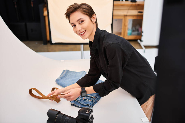 радісна жінка-фотограф готується робити фотографії джинсів і поясів і посміхається на камеру - Фото, зображення