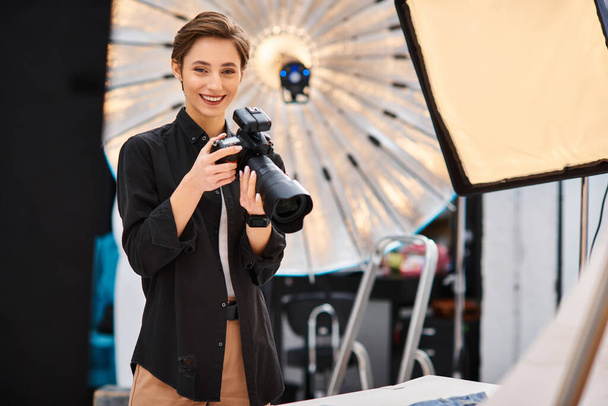radosna atrakcyjna kobieta w luźnym stroju uśmiechnięta do aparatu fotograficznego i trzymająca kamerę w swoim studiu fotograficznym - Zdjęcie, obraz