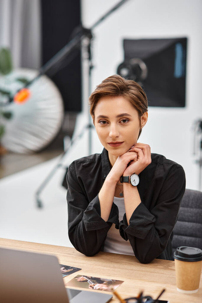 jeune femme attrayante en tenue décontractée assis au bureau dans son studio de photo et regardant la caméra - Photo, image