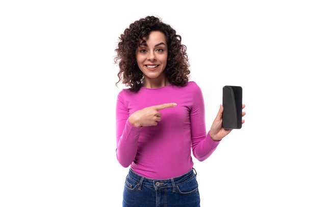 jeune fashionista bouclée dame brune vêtue d'un col roulé couleur lilas frais montre l'écran d'un smartphone. - Photo, image