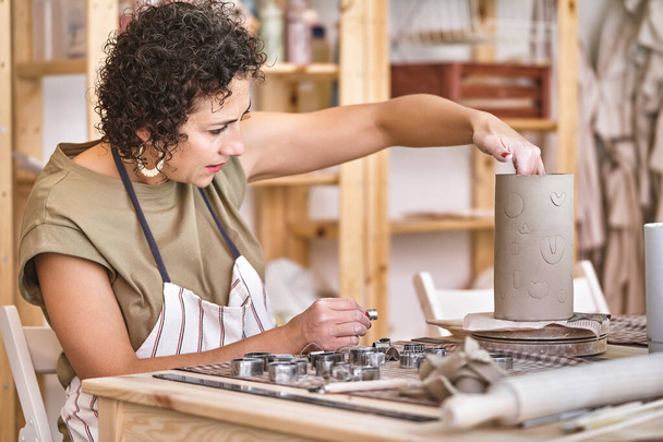 Konzentrierter Töpfer in gestreifter Schürze trägt in einer Werkstatt kreative Abdrücke auf ein zylindrisches Tonstück auf - Foto, Bild