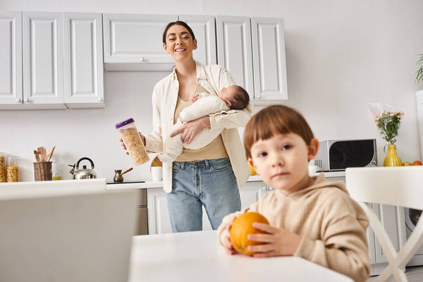 vrolijk moeder in casual kleding houden cornflakes in de buurt van haar peuter zoon terwijl het houden van haar pasgeboren - Foto, afbeelding