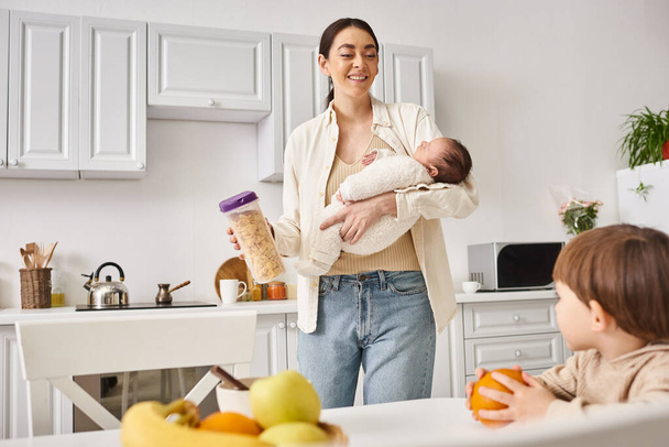 szczęśliwa matka w stroju casual trzymając płatki kukurydziane w pobliżu jej syna malucha podczas trzymania jej noworodka - Zdjęcie, obraz