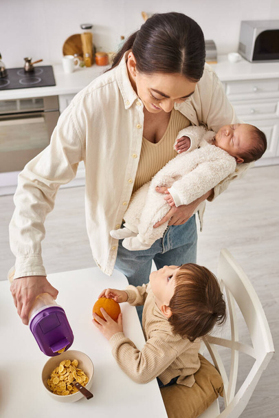 feliz hermosa mujer vertiendo copos de maíz a su adorable hijo pequeño mientras sostiene a su bebé recién nacido - Foto, Imagen