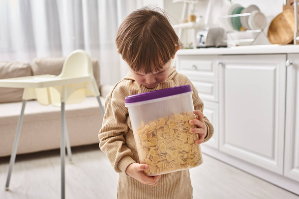 adorable maluch chłopiec w casual homewear gospodarstwa opakowanie płatków kukurydzianych podczas śniadania w kuchni - Zdjęcie, obraz