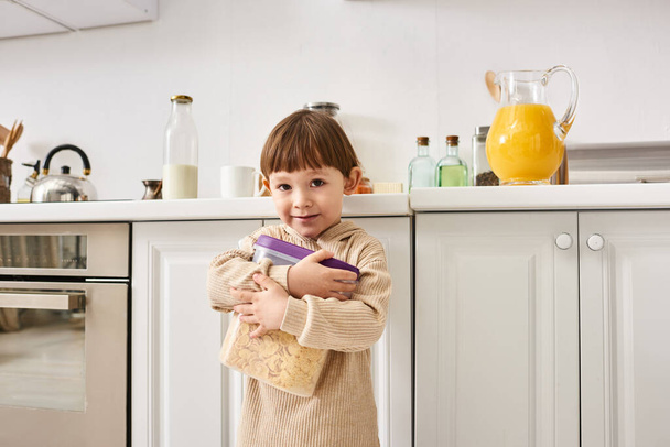 adorable niño en cómoda ropa de casa sosteniendo paquete de copos de maíz durante el desayuno en la cocina - Foto, imagen