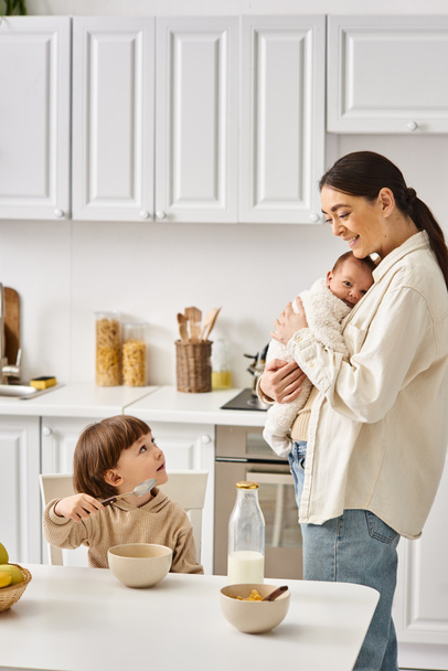 радісна приваблива мати позує поруч зі своїм маленьким сином з новонародженим в руках під час сніданку - Фото, зображення