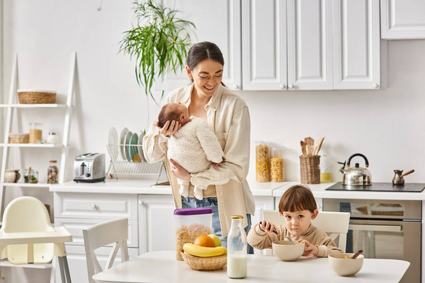 vrolijk aantrekkelijke moeder poseren naast haar peuter zoon met pasgeboren in handen tijdens het ontbijt - Foto, afbeelding