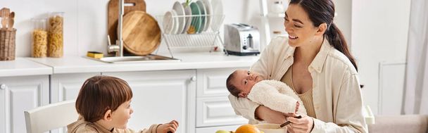Весело красивая мать позирует рядом со своим малышом сыном с новорожденным в руках во время завтрака, баннер - Фото, изображение