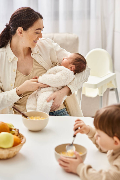στοργική όμορφη μητέρα παίρνει πρωινό με το μικρό της γιο και κρατώντας το νεογέννητο μωρό της - Φωτογραφία, εικόνα