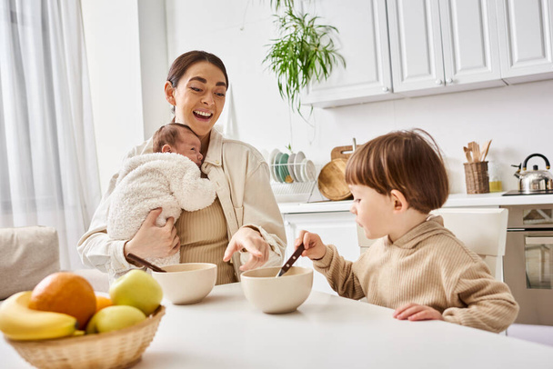 opiekuńcza atrakcyjna matka jedząca śniadanie ze swoim synkiem i trzymająca swoje nowonarodzone dziecko - Zdjęcie, obraz
