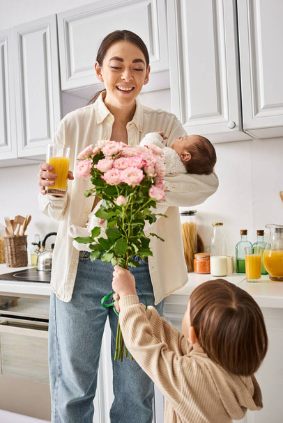adorable toddler boy giving flowers to his attractive merry mother with his newborn brother in hands - Photo, Image