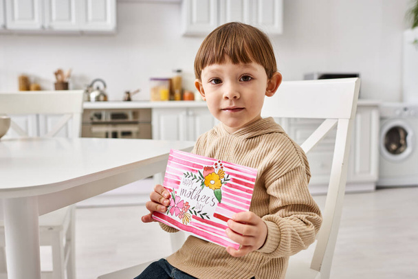 очаровательный маленький мальчик в удобной одежде сидит и держит поздравительную открытку на День матери - Фото, изображение
