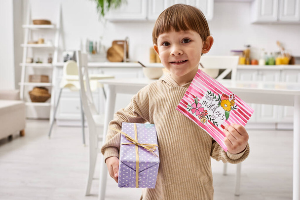 очаровательный милый малыш в удобной домашней одежде позирует с подарком и поздравительной открыткой на День Матери - Фото, изображение