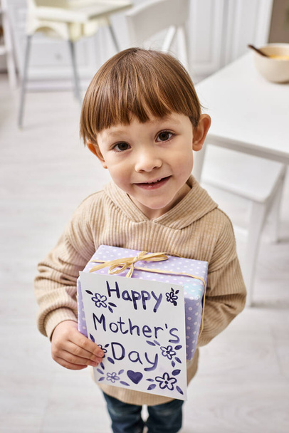 αξιολάτρευτο χαριτωμένο αγοράκι σε casual homewear ποζάρουν με το παρόν και ευχετήρια κάρτα για την Ημέρα της Μητέρας - Φωτογραφία, εικόνα