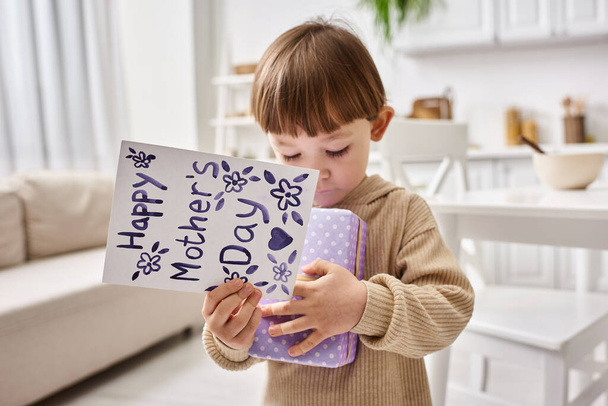 очаровательный милый маленький мальчик в удобной домашней одежде позирует с подарком и поздравительной открыткой на День матери - Фото, изображение