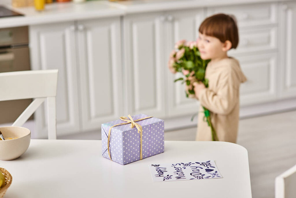 adorable niño en ropa de casa con ramo de flores cerca de la mesa con regalo y postal en ella - Foto, Imagen