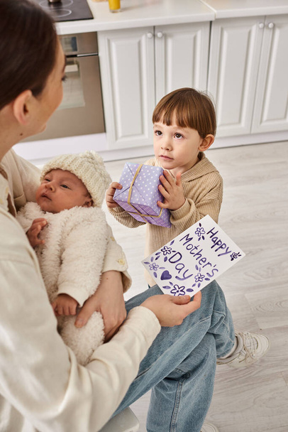 adorable boy giving gift and postcard to his mom while she holding his newborn brother, Mothers day - Photo, Image