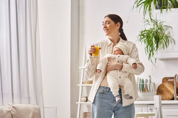 feliz madre amorosa sosteniendo a su bebé recién nacido y sosteniendo jugo de naranja, crianza moderna - Foto, imagen