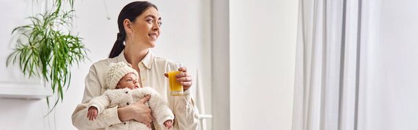 χαρούμενη στοργική μητέρα κρατώντας το νεογέννητο αγοράκι της και κρατώντας χυμό πορτοκάλι, σύγχρονη ανατροφή των παιδιών, λάβαρο - Φωτογραφία, εικόνα