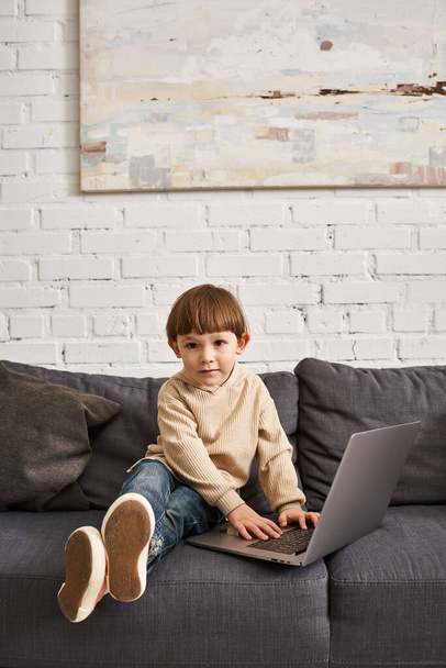 чарівний милий хлопчик-малюк в затишному домашньому одязі, сидячи на дивані з ноутбуком і дивлячись на камеру - Фото, зображення