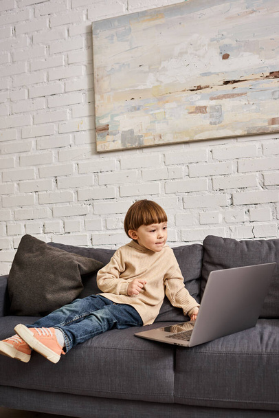 συμπυκνωμένο αξιολάτρευτο χαριτωμένο αγοράκι σε ζεστό homewear κάθεται στον καναπέ και κοιτάζοντας το laptop - Φωτογραφία, εικόνα