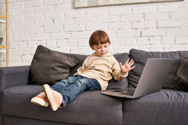 εστιασμένη αξιολάτρευτο χαριτωμένο αγοράκι σε άνετο homewear κάθεται στον καναπέ και κοιτάζοντας το laptop - Φωτογραφία, εικόνα