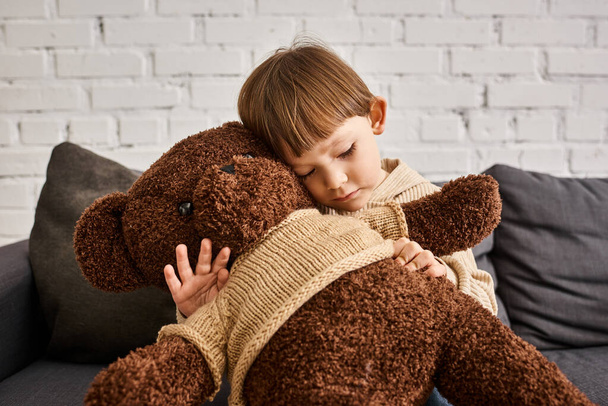 cute little boy in casual warm homewear hugging his teddy bear while sitting on couch at home - Photo, Image