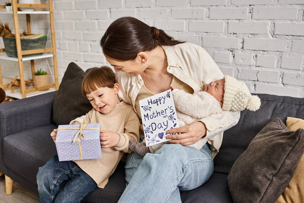 gyönyörű nő ül a kanapén a fiaival ajándékkal és üdvözlőkártyával anyák napján - Fotó, kép