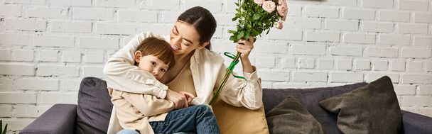 schöne Mutter posiert auf dem Sofa mit ihrem niedlichen Kleinkind Sohn mit Blumenstrauß, Muttertag, Banner - Foto, Bild