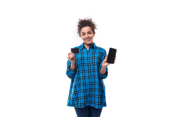 jovem bela mulher europeia com cabelo encaracolado vestido com uma camisa xadrez azul detém um cartão de desconto e um smartphone. - Foto, Imagem