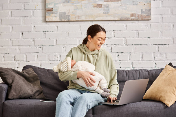 goed uitziende vrolijke moeder in casual kleding met videocall en het houden van haar pasgeboren baby jongen - Foto, afbeelding