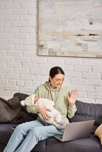 goed uitziende vrolijke moeder in casual kleding met videocall en het houden van haar pasgeboren baby jongen - Foto, afbeelding