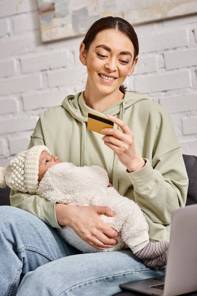 beautiful smiling mother in homewear holding her baby boy in font of laptop with credit card in hand - Photo, Image