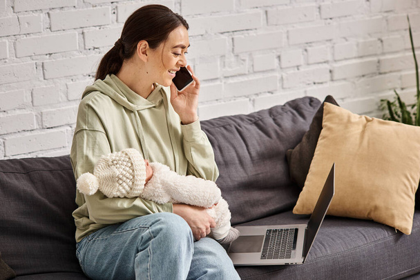 весела приваблива жінка в затишному вбранні дивиться на ноутбук і тримає дитину під час розмови по телефону - Фото, зображення