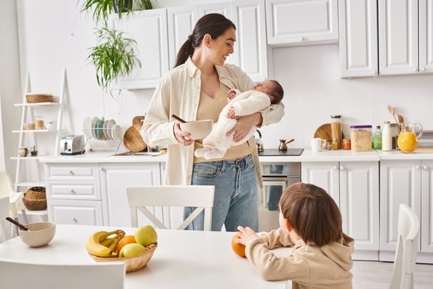 gioiosa donna attraente preparare la colazione per il suo bambino figlio mentre tiene il suo neonato ragazzo - Foto, immagini