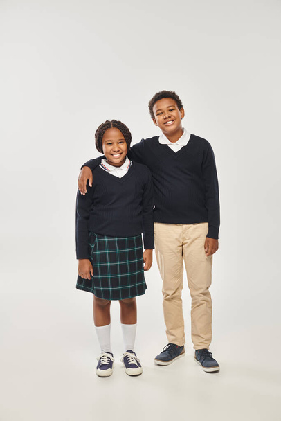 happy african american boy in school uniform hugging girl in plaid skirt while standing on grey - Photo, Image
