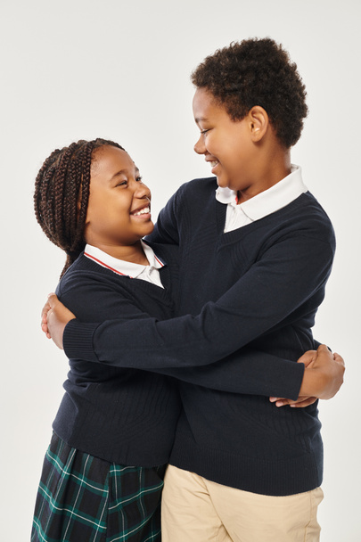 vrolijk tiener afrikaanse amerikaanse kinderen in school uniform knuffelen elkaar op grijze achtergrond - Foto, afbeelding