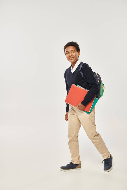 heureux écolier afro-américain en uniforme tenant des cahiers et debout sur fond gris - Photo, image
