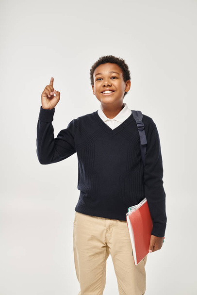 szczęśliwy afrykański amerykański uczeń w mundurze trzymając zeszyty i pokazując pomysł na szarym tle - Zdjęcie, obraz