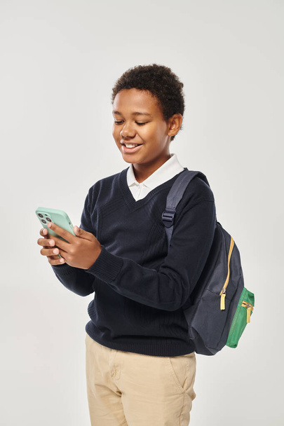 heureux écolier afro-américain en uniforme en utilisant smartphone et debout sur fond gris - Photo, image
