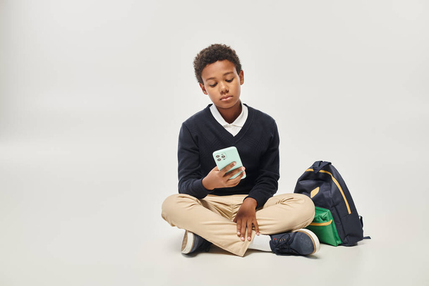 écolier afro-américain en uniforme à l'aide d'un smartphone et assis près du sac à dos sur fond gris - Photo, image