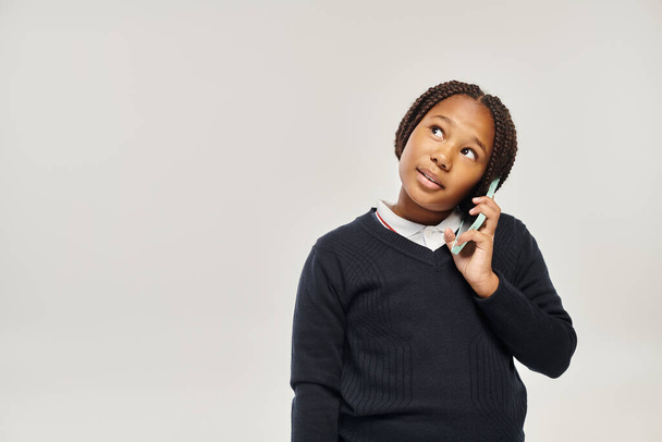 preadolescente afroamericana colegiala en uniforme hablando en teléfono inteligente sobre fondo gris, mirar hacia otro lado - Foto, imagen