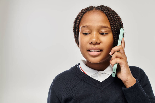 preteen african american schoolgirl in uniform talking on smartphone on grey background, look down - Photo, Image