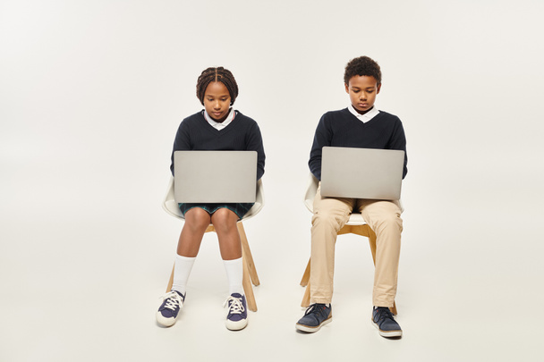 skoncentrowane afrykańskie amerykańskie dzieci w mundurach, korzystające z laptopów i siedzące na szarym tle - Zdjęcie, obraz