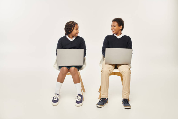 веселі афроамериканські школярі в уніформі з використанням ноутбуків і сидячи на сірому тлі - Фото, зображення