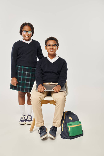 весела афроамериканська дівчина в уніформі, що стоїть біля школяра з ноутбуком на сірому фоні - Фото, зображення