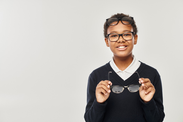 optimista afroamericano chico en suéter y gafas sosteniendo gafas y mirando a la cámara en gris - Foto, imagen