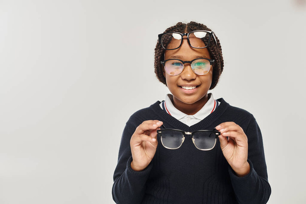Χαρούμενη Αφροαμερικάνα με πουλόβερ και γυαλιά να κρατάει γυαλιά και να κοιτάει την κάμερα στο γκρι - Φωτογραφία, εικόνα