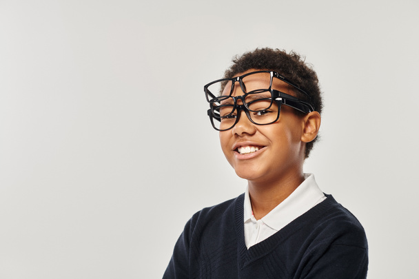 optimistic african american boy in sweater and eyewear holding glasses and looking at camera on grey - Photo, Image